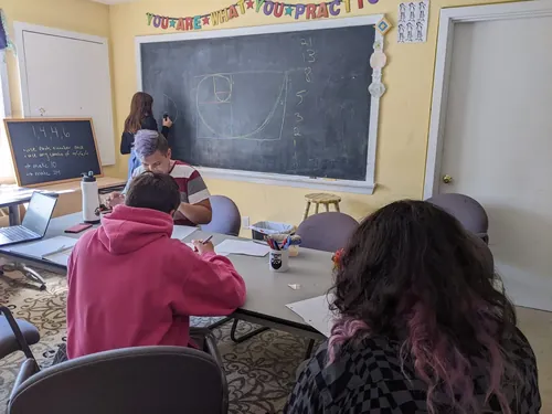 A member works on a Fibonacci spiral at the chalkboard while others work around a table