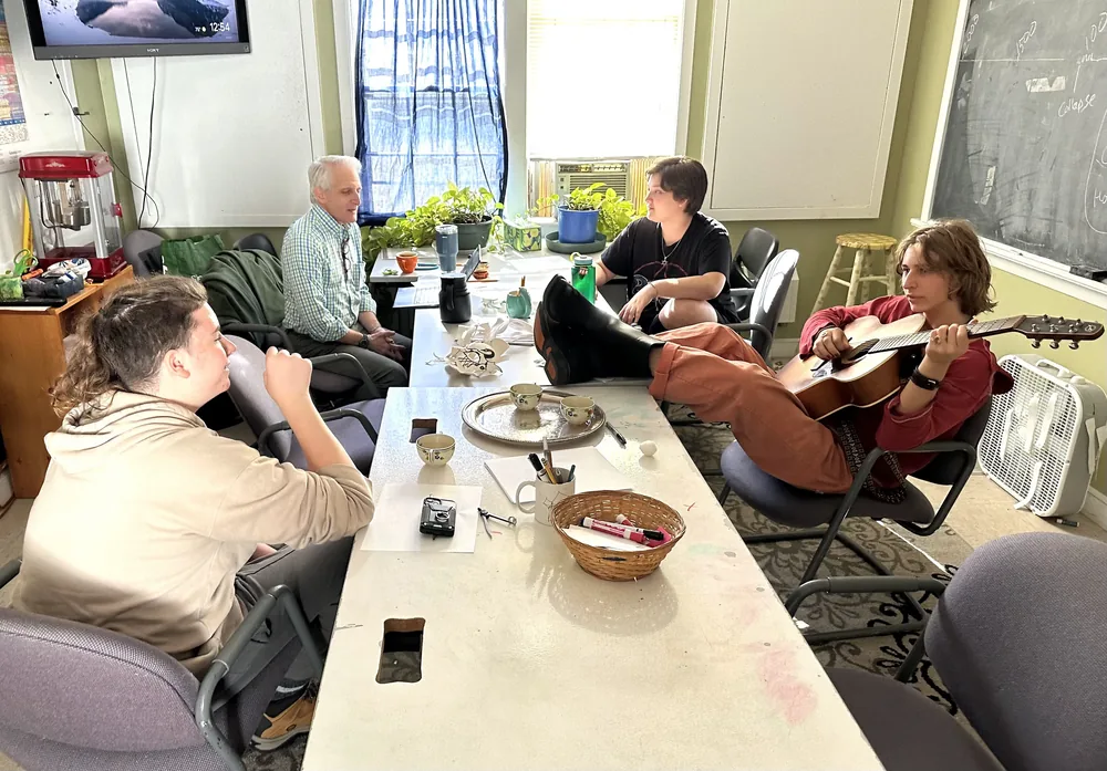 Three members and one teacher sit around a table during a seminar. One member plays a guitar during the conversation.